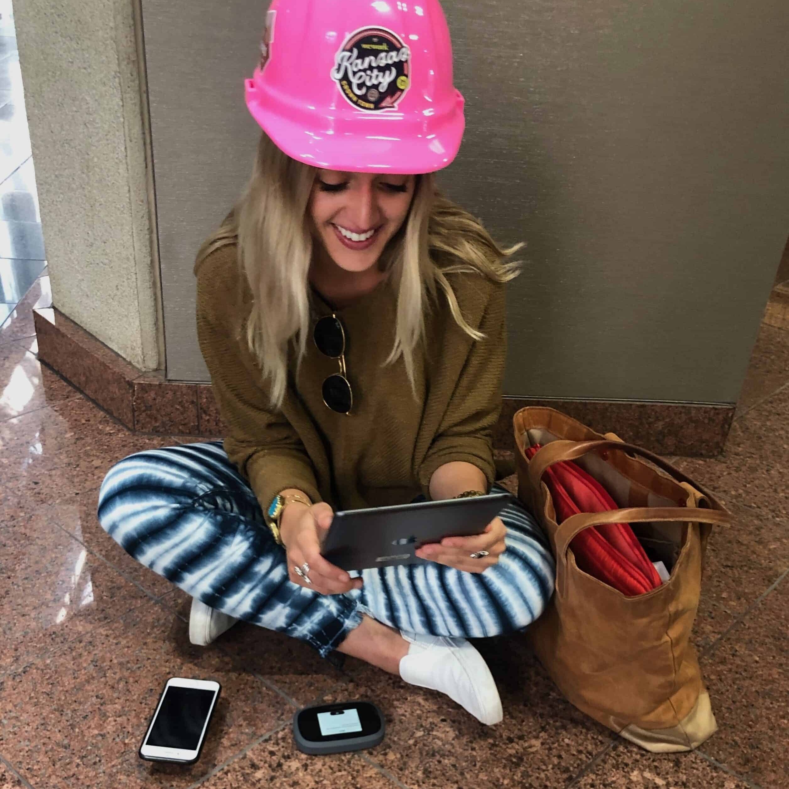 A woman wearing a pink hard hat sits on the floor using a tablet, with a smartphone and a brown bag beside her.